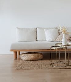 A chic minimalist living room featuring a white sofa with cushions, dried flowers in a vase, and a wooden floor.
